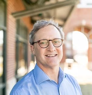 Smiling man in glasses and a light blue shirt standing outdoors near brick arches