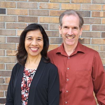Two smiling people standing in front of a brick wall, one in a black jacket and patterned blouse, one in a red shirt
