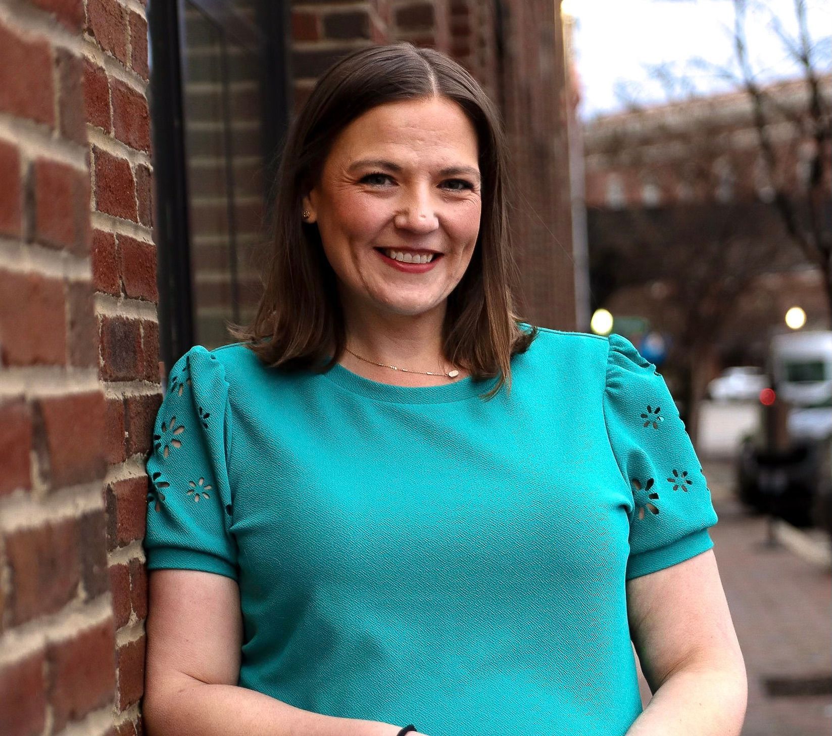Woman in a turquoise shirt smiling beside a brick wall on a city sidewalk