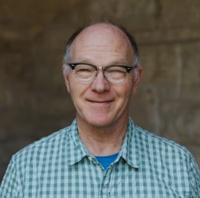 Smiling man in glasses wearing a green plaid shirt against a blurred brown background