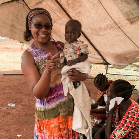 Smiling woman holding a child under a tent, with two people seated nearby.