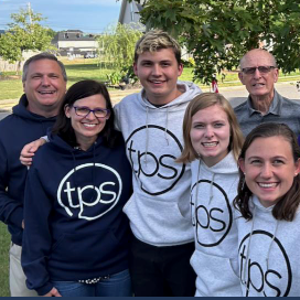Five smiling people outdoors in TPS hoodies, standing together in a group selfie.