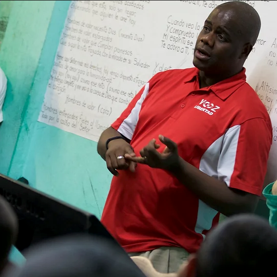 Man in red polo speaking to a group in a classroom, gesturing beside a whiteboard.