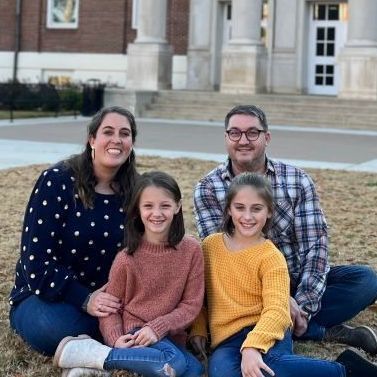 Four people sitting on grass in front of a building, smiling at the camera.