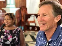 Smiling man in blue plaid shirt in foreground, woman seated behind him in a church-like interior