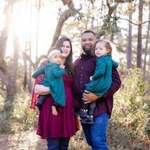 Family of four standing outdoors in a wooded area, holding two small children.