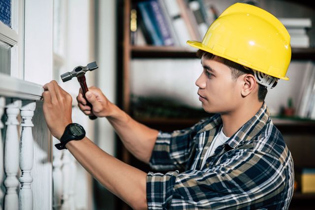 Worker in a yellow hard hat hammering a window frame indoors