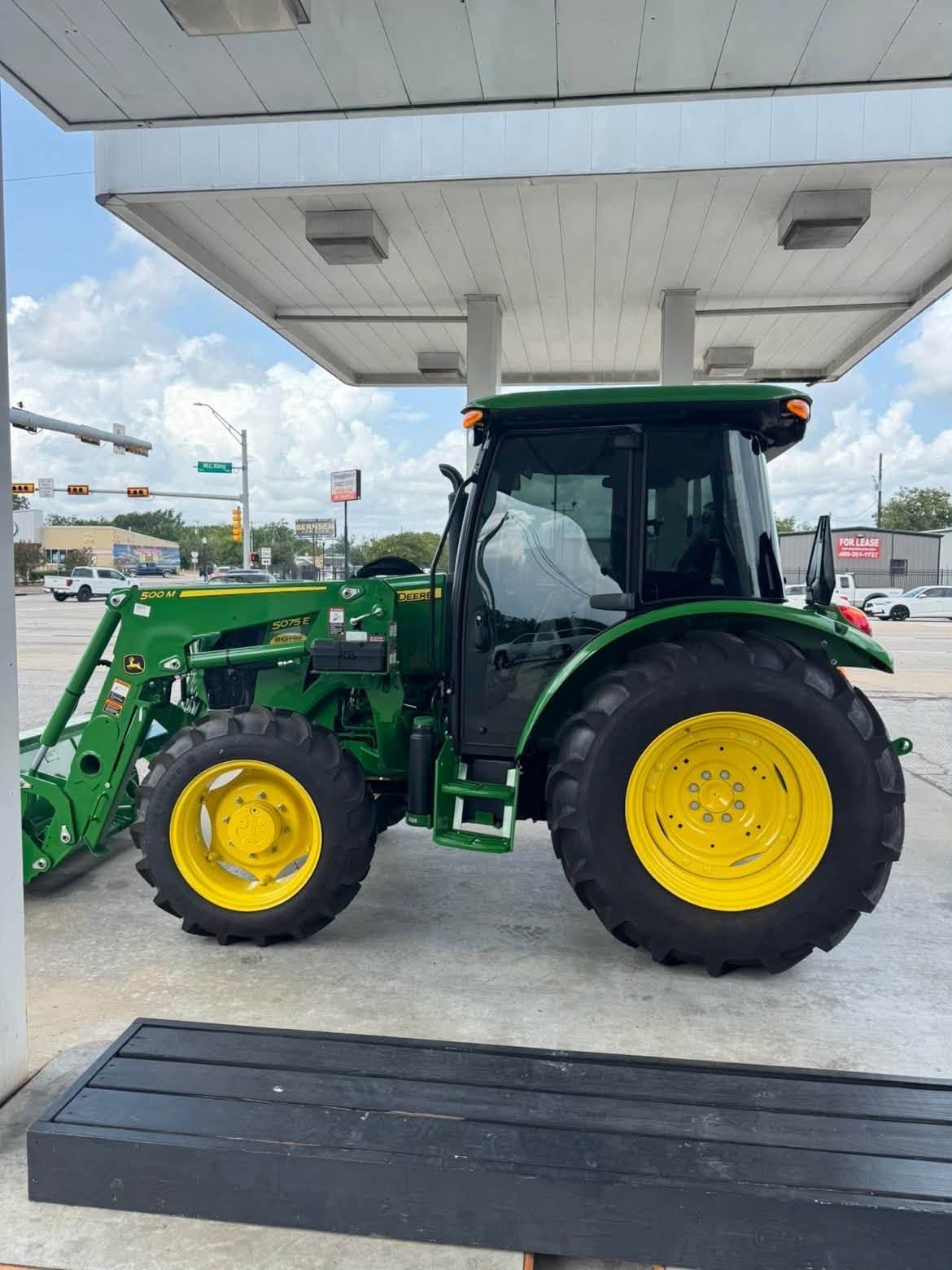 Green tractor with yellow wheels parked under a gas station canopy near a road
