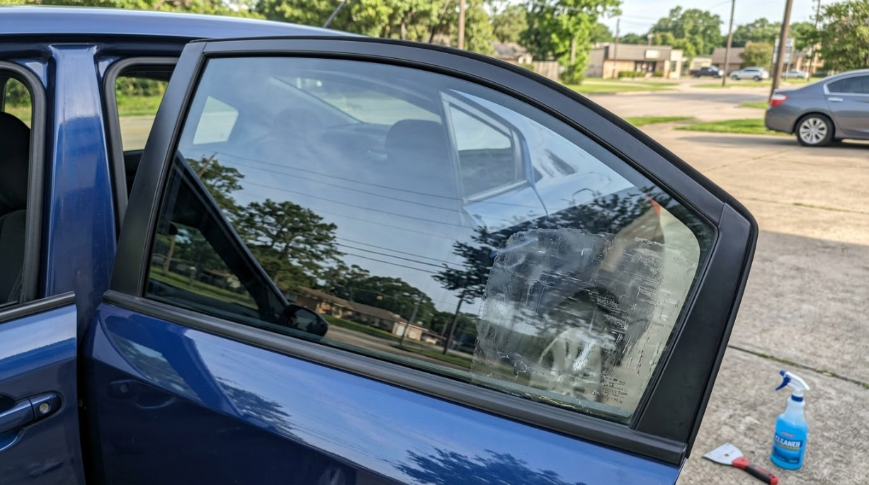 Blue car with open door and shattered side window, parked beside a road.