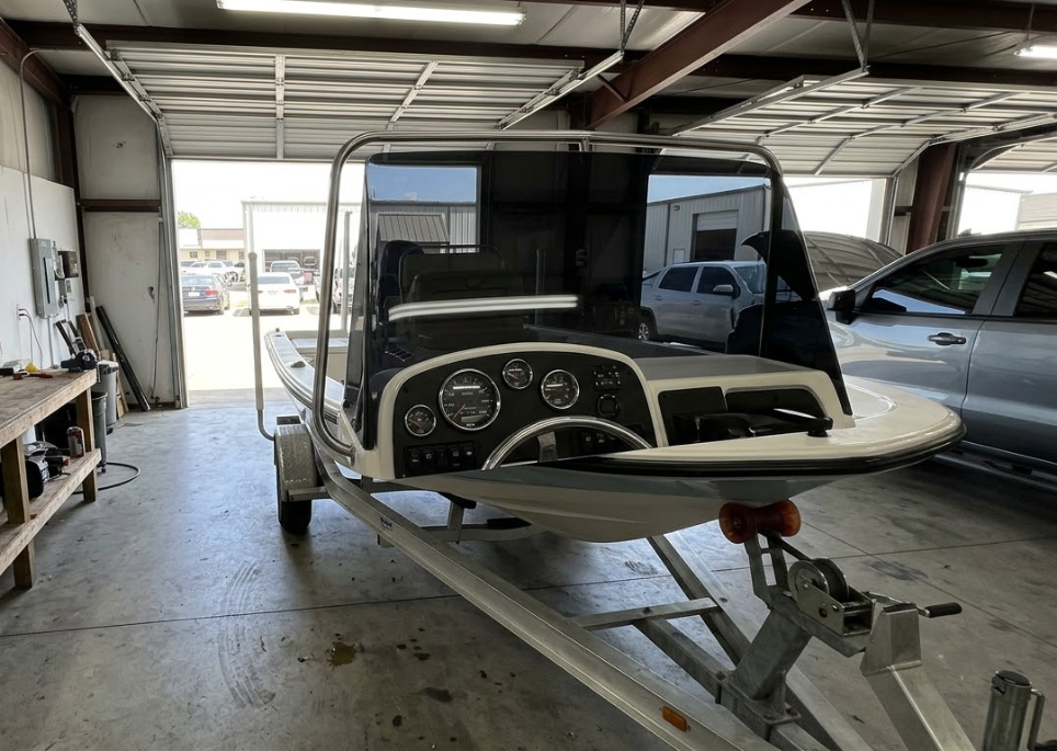 Small boat on a trailer inside a workshop, with dashboard visible and another boat in the background.