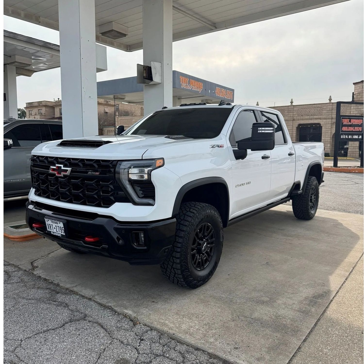 White Chevrolet pickup truck with black off-road tires parked at a gas station under a canopy.