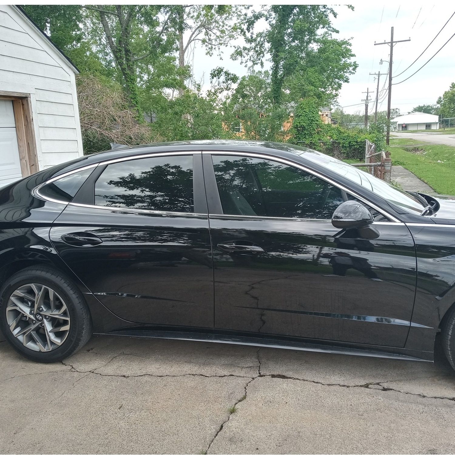 Black sedan parked beside a white building on a cracked driveway, with trees and power lines behind it