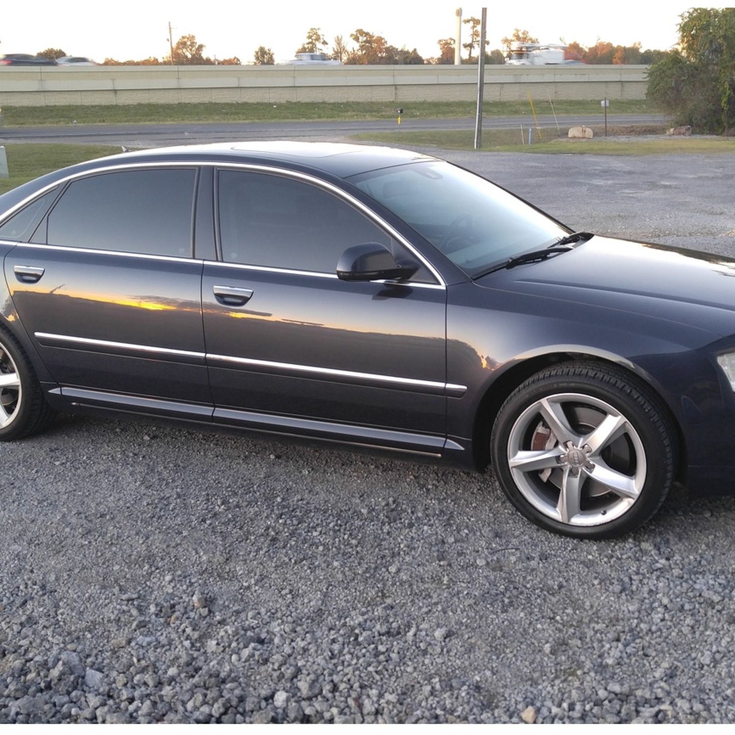 Dark gray sedan parked on gravel beside a road at sunset
