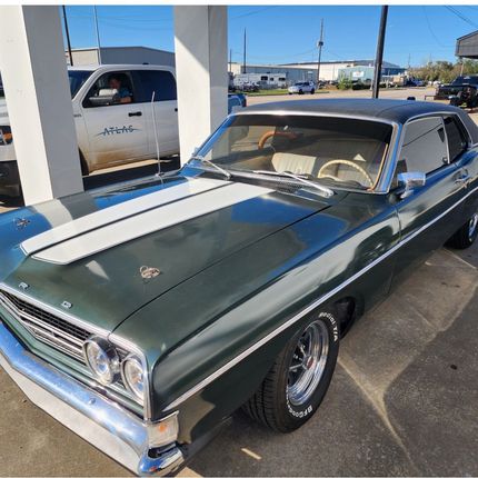 Classic dark green muscle car with white racing stripes parked at a gas station