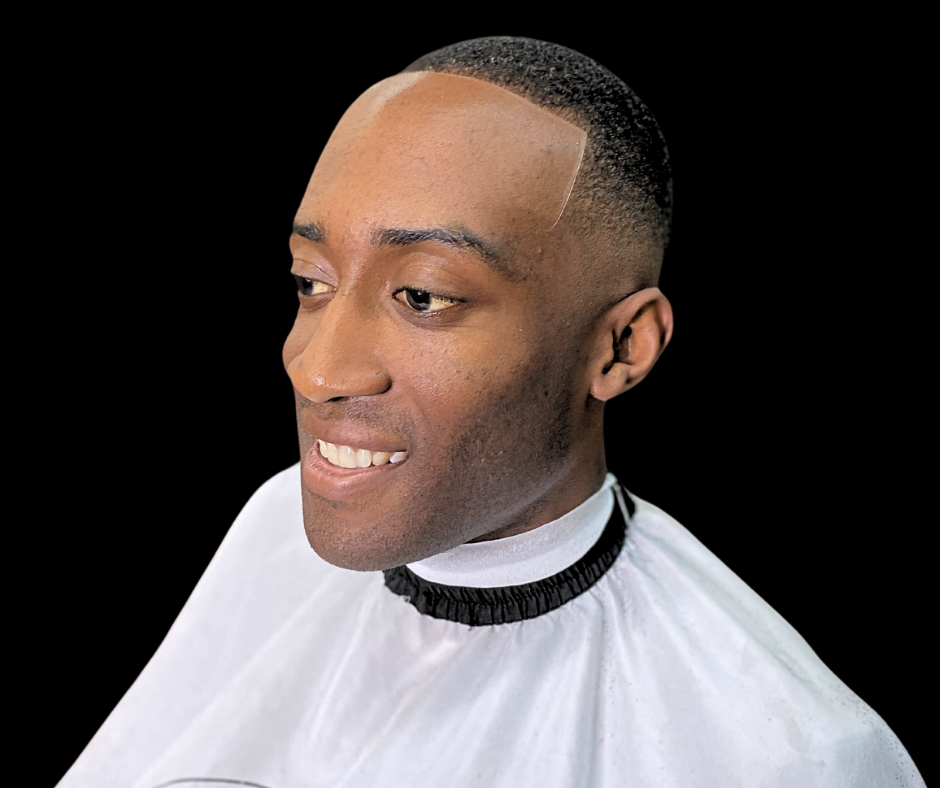 Man with a high-fade haircut smiles while wearing a barber's cape against a black background.