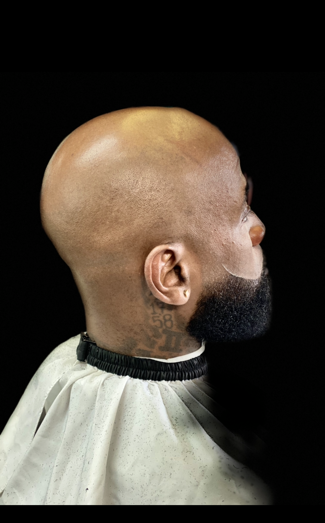 Side profile of a person with a shaved head and a trimmed beard, wearing a barber cape against a black background.