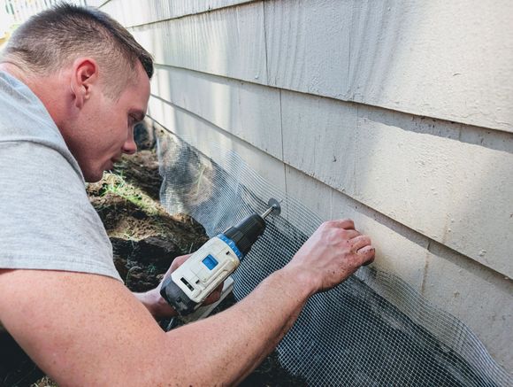Man using a drill to install mesh along a wall's siding near the ground outdoors.