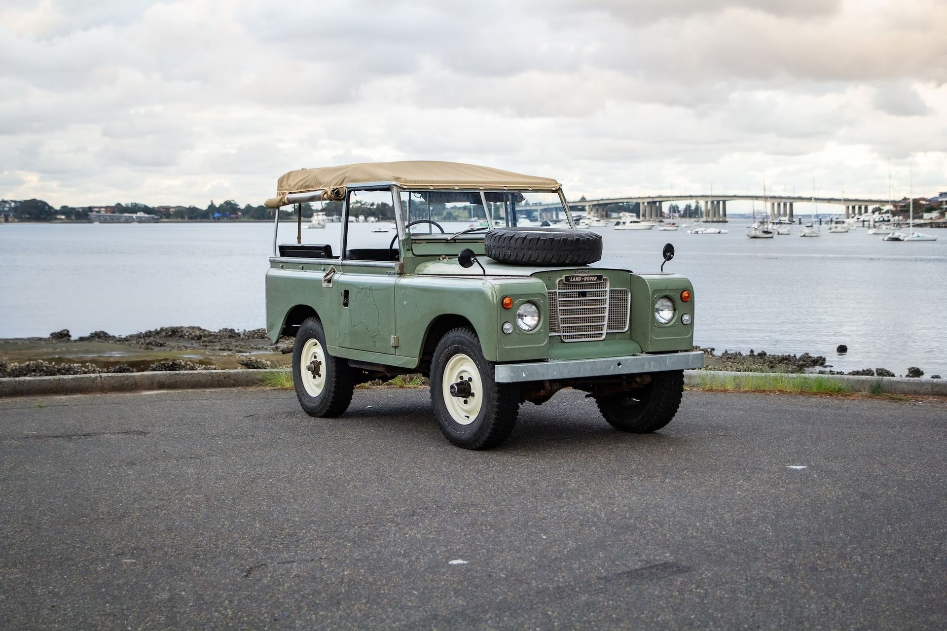 Land Rover vintage verte garée près de l'eau. Toit beige, roue de secours sur le capot, jantes blanches, ciel couvert.