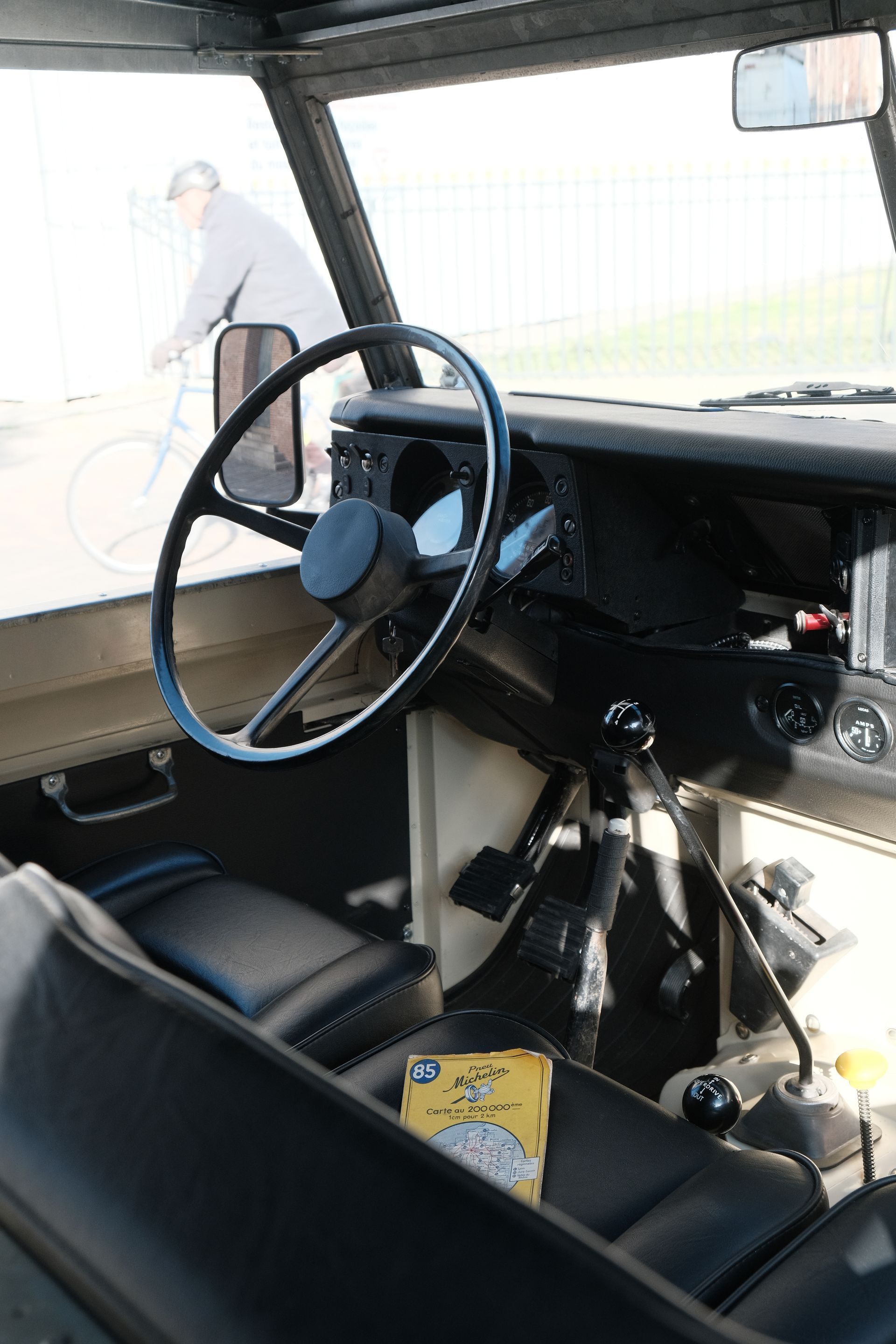 Intérieur d'un Land Rover : volant noir, levier de vitesses, tableau de bord et un cycliste aperçu à travers le pare-brise.
