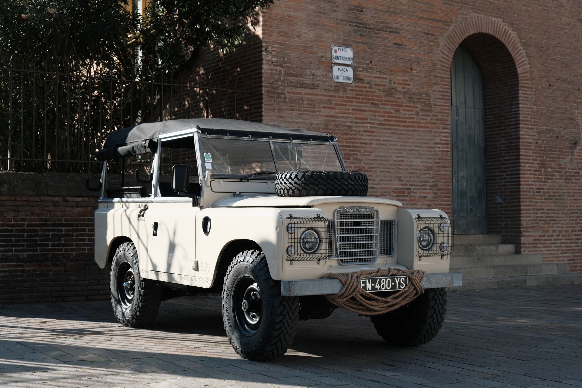 Land Rover beige garée devant un bâtiment en briques. Roue de secours sur le capot, jantes noires et corde enroulée sur le pare-chocs.