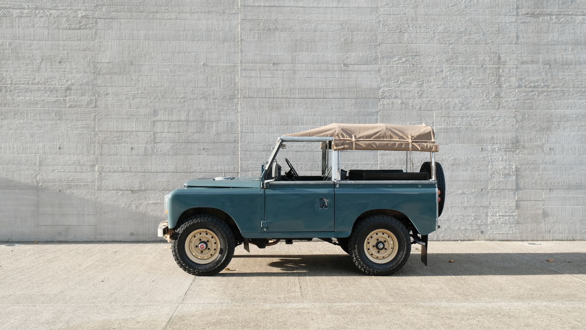 Land Rover vintage bleue avec toit et roues beiges, garée devant un mur en béton.
