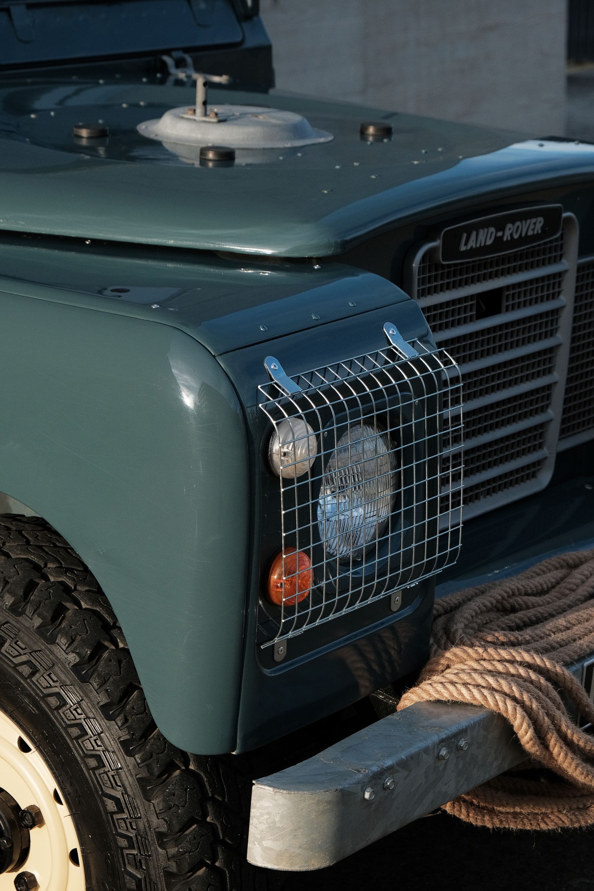 Gros plan sur un Land Rover Defender turquoise avec calandre de phares et corde enroulée sur le pare-chocs.
