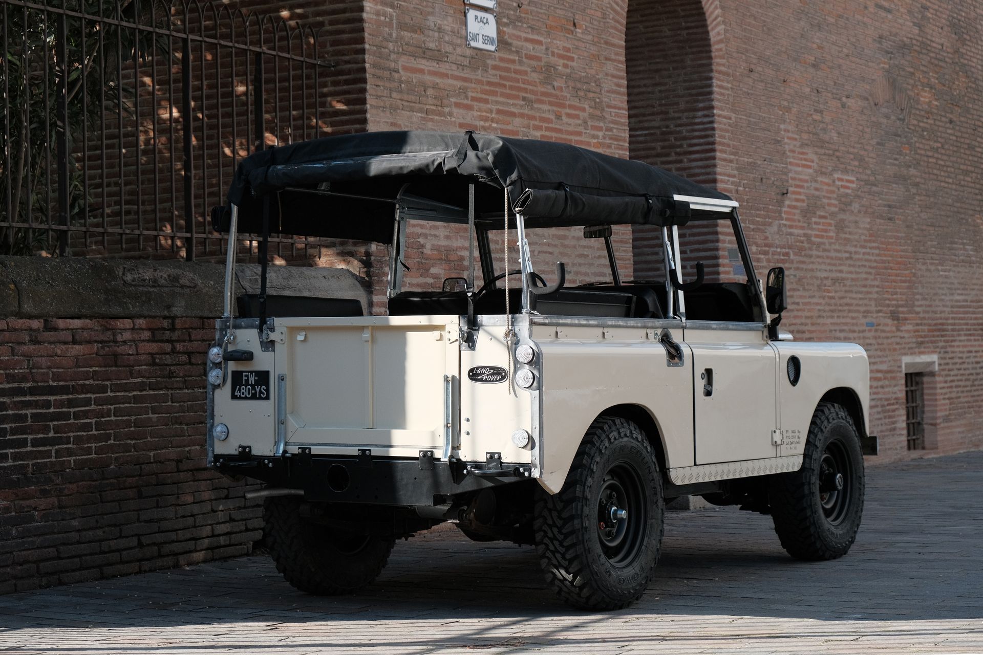 Land Rover Defender beige, capote souple noire, garé à côté d'un mur de briques.