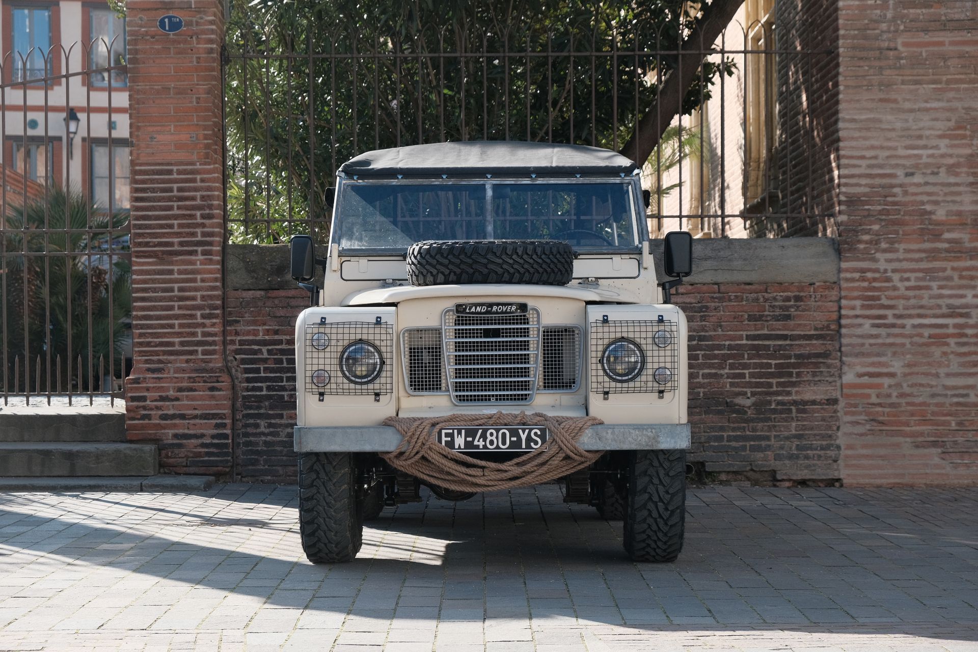 Land Rover beige garée devant un bâtiment en briques. Roue de secours sur le capot, pare-chocs en corde.