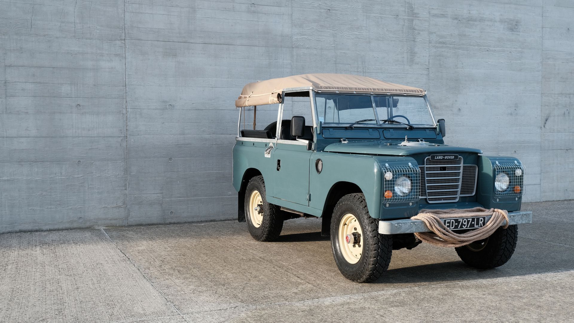 Un Land Rover vintage bleu avec un toit beige est garé contre un mur en béton gris.