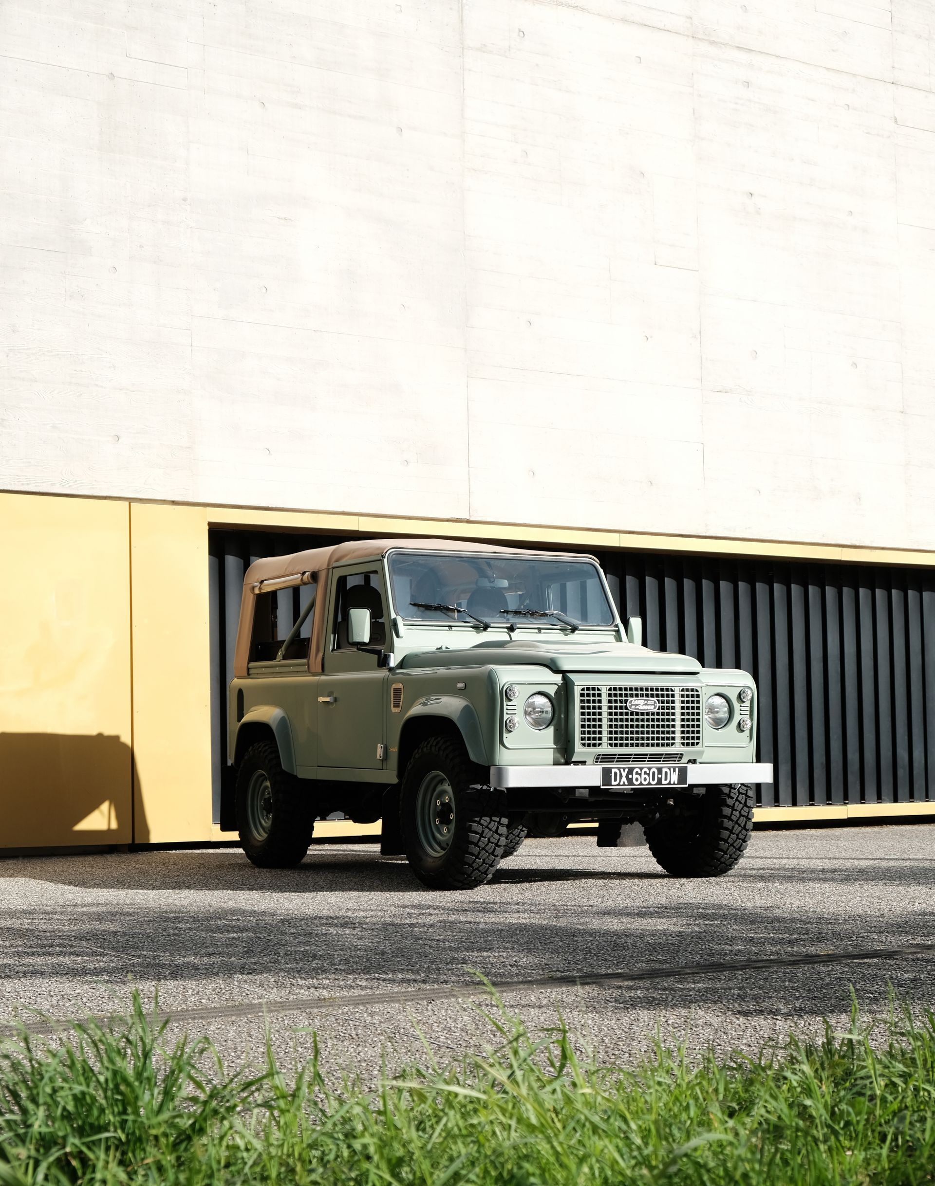 Un defender 90 VERT 1CD TD4 soft top beige, bâche stayfast ExmoorTrim , édition héritage garé devant un bâtiment en béton banché brut.