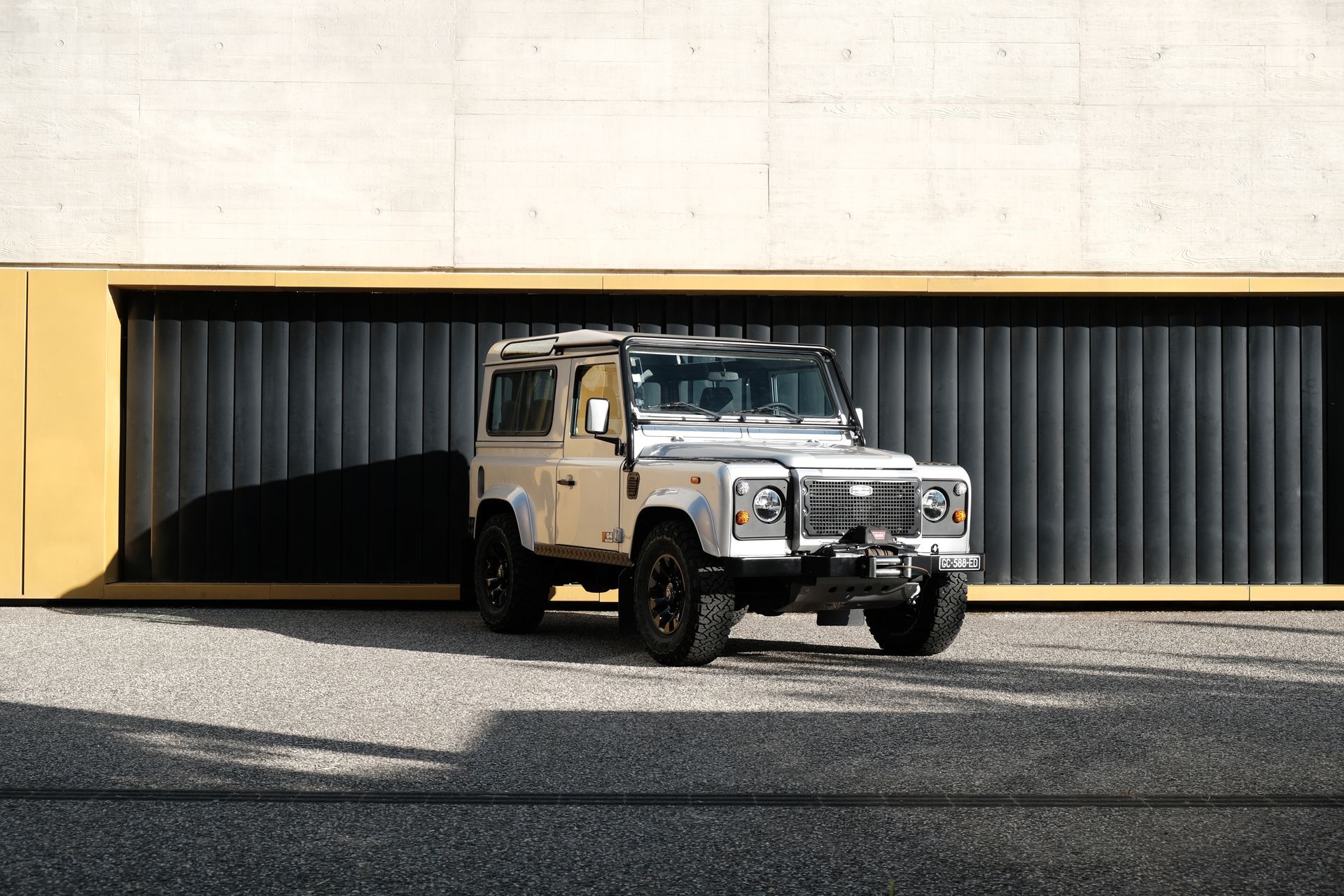 Un Land Rover Defender vert et blanc roule sur une route côtière, surplombant une plage et des collines verdoyantes.