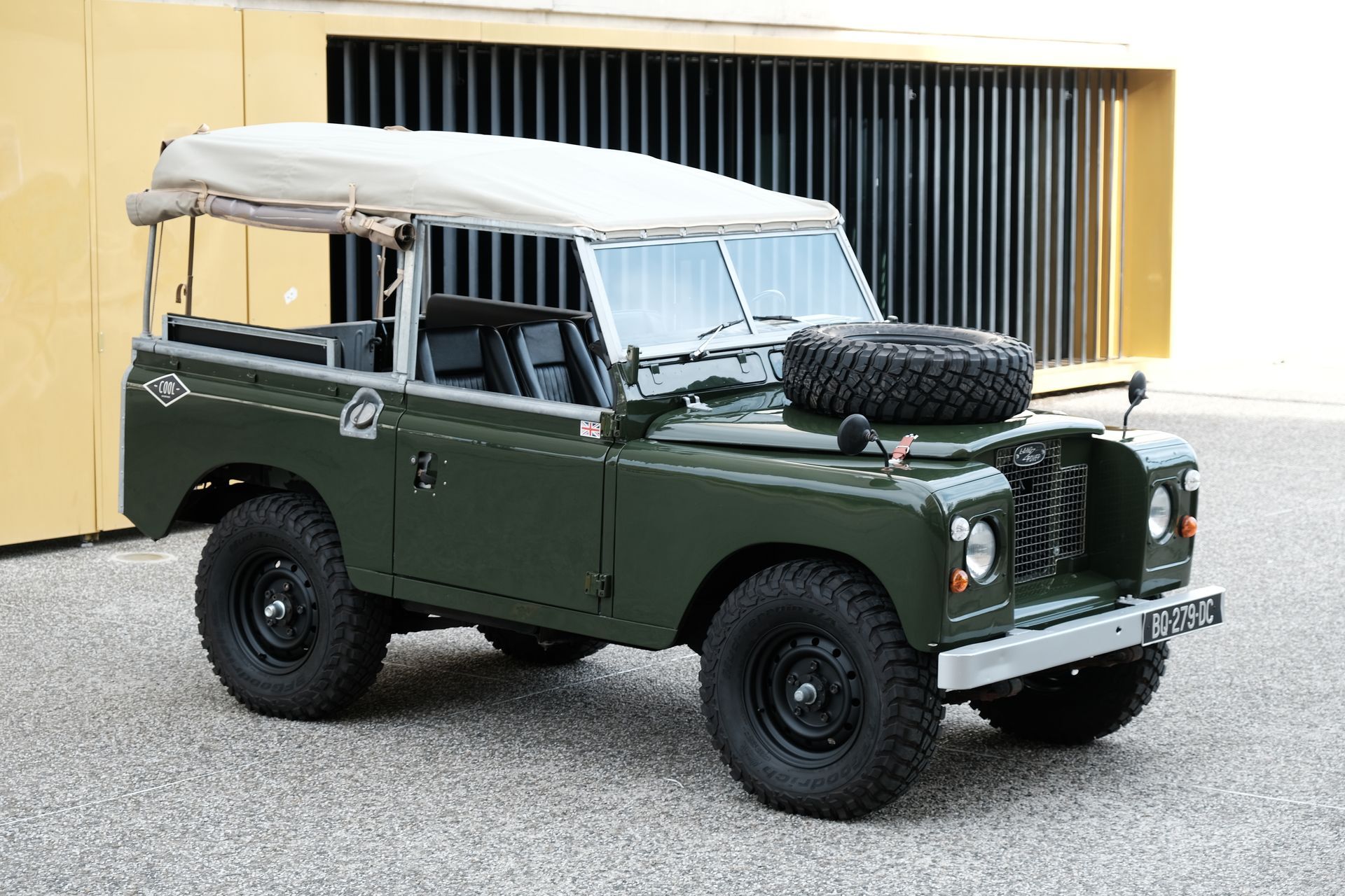 Land Rover 88 Bronze green, véhicule tout-terrain avec toit soft top beige, roue de secours sur le capot, garée à l'extérieur.