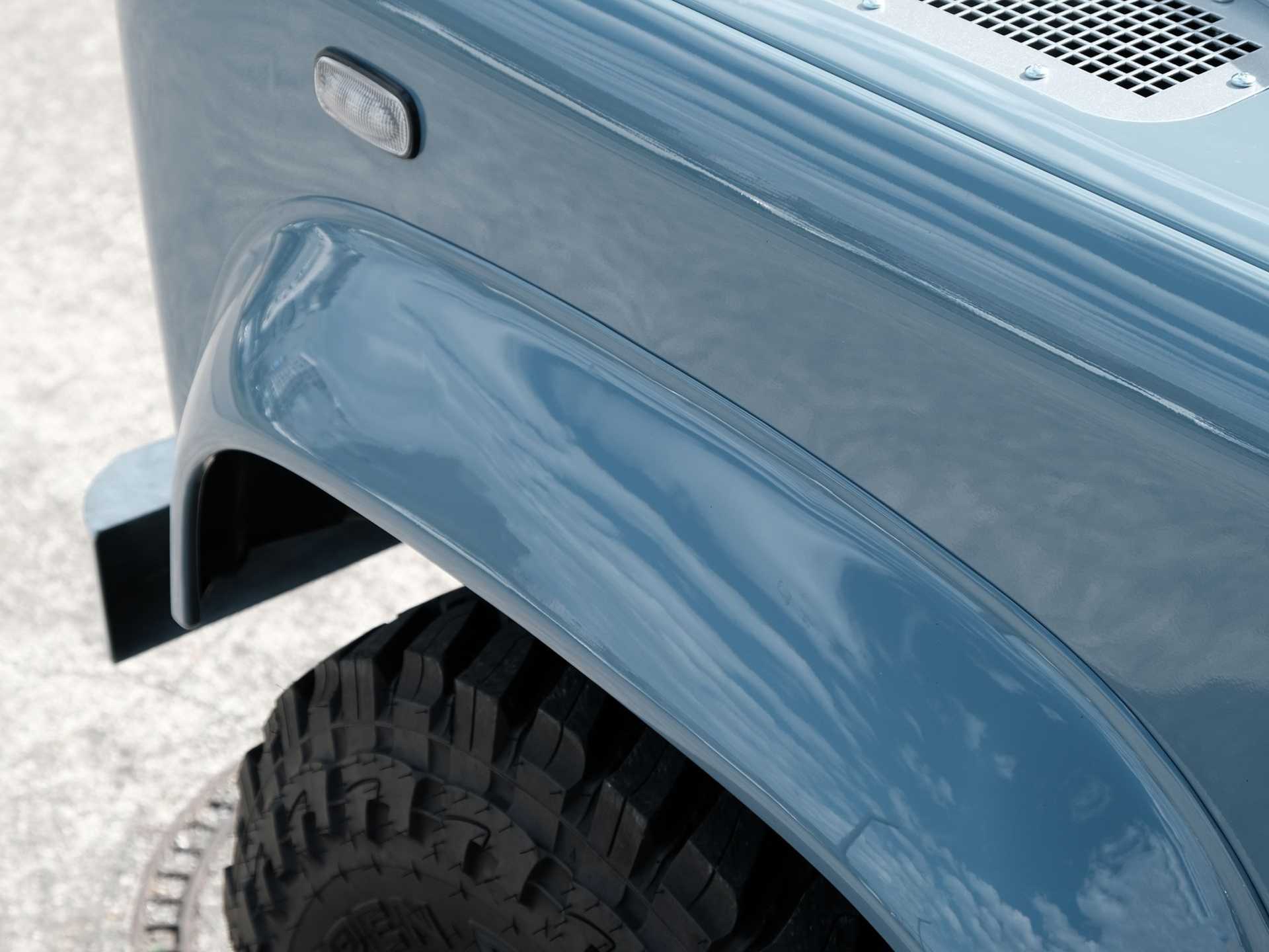 Land Rover Defender bleu roulant sur une plage de sable, capote souple beige, ciel couvert.
