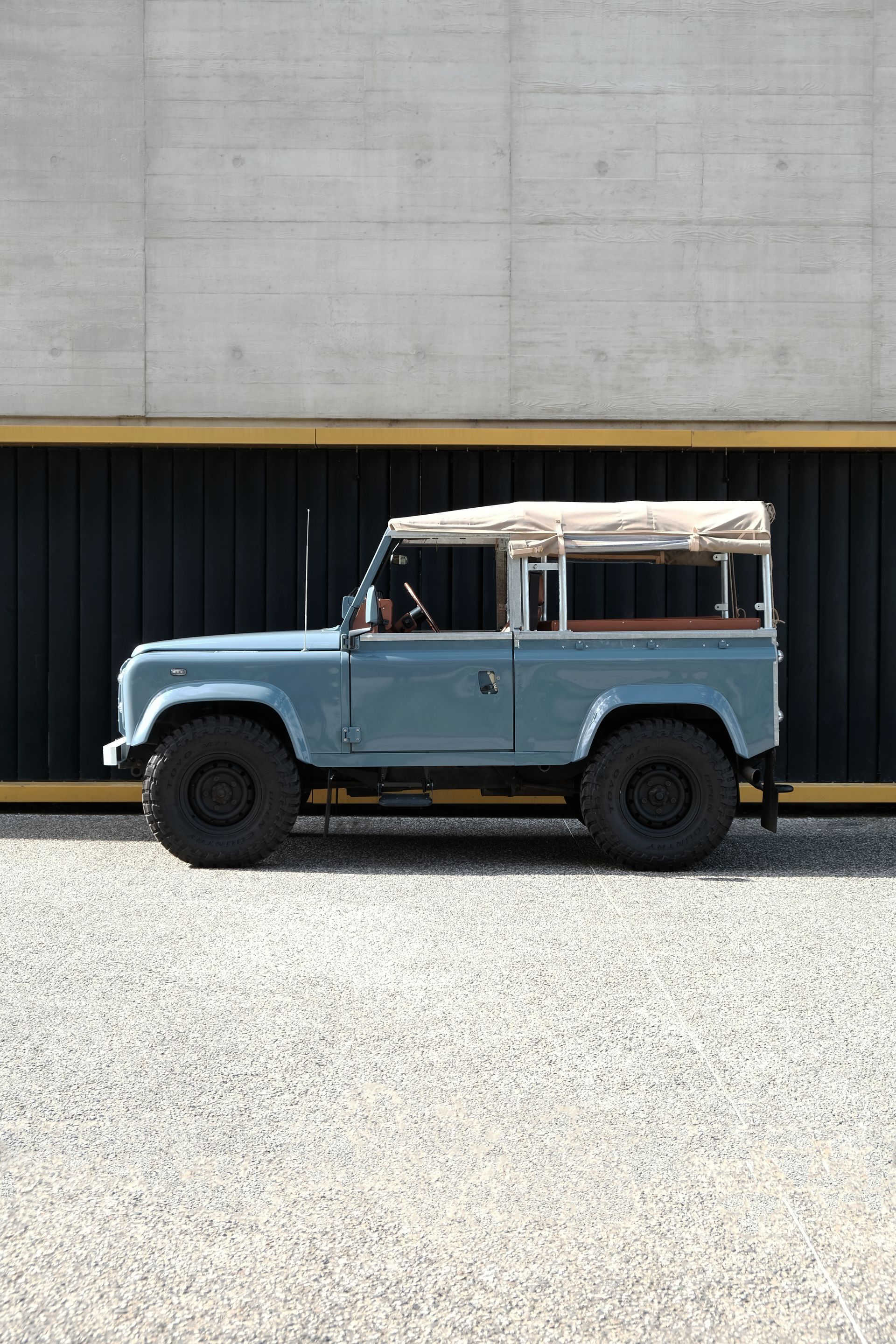Un Land Rover Defender bleu clair avec un toit beige est garé sur du gravier, devant un bâtiment en béton.
