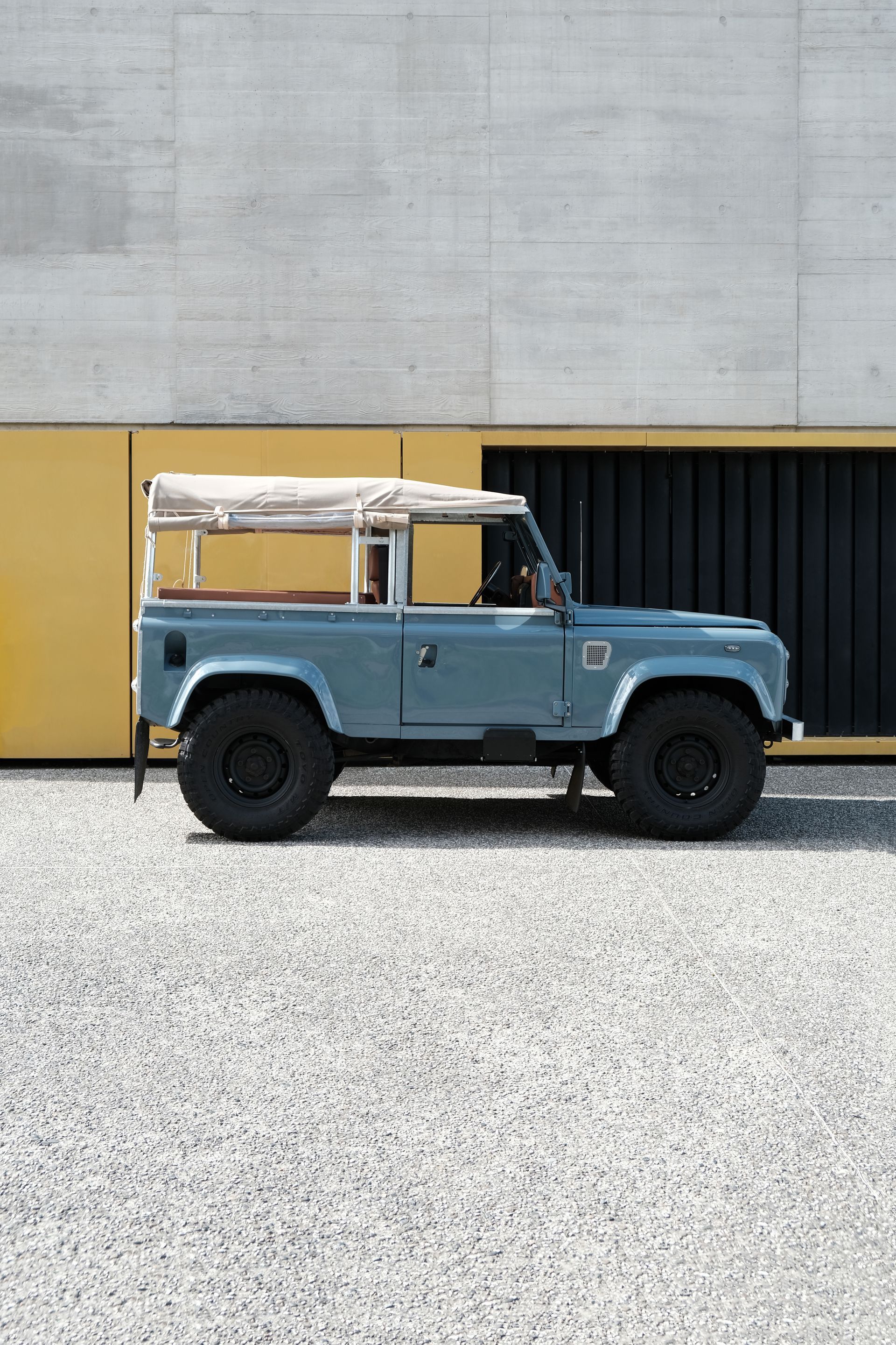 Un Land Rover Defender bleu avec un toit beige, garé sur du gravier devant un bâtiment jaune et gris.
