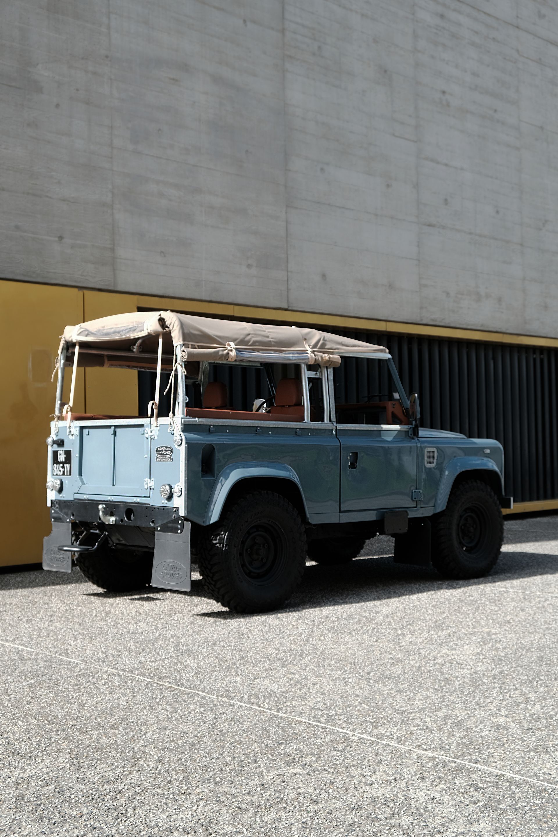 Un Land Rover Defender bleu clair avec une capote souple beige est garé contre un mur en béton texturé.