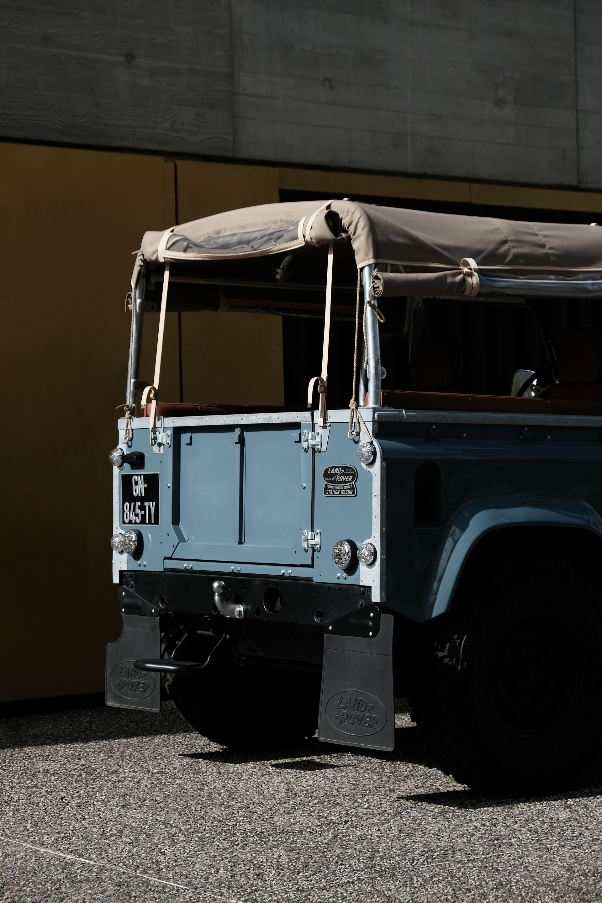 Un Land Rover Defender bleu clair avec une capote en toile, garé sur du gravier à côté d'un mur jaune.