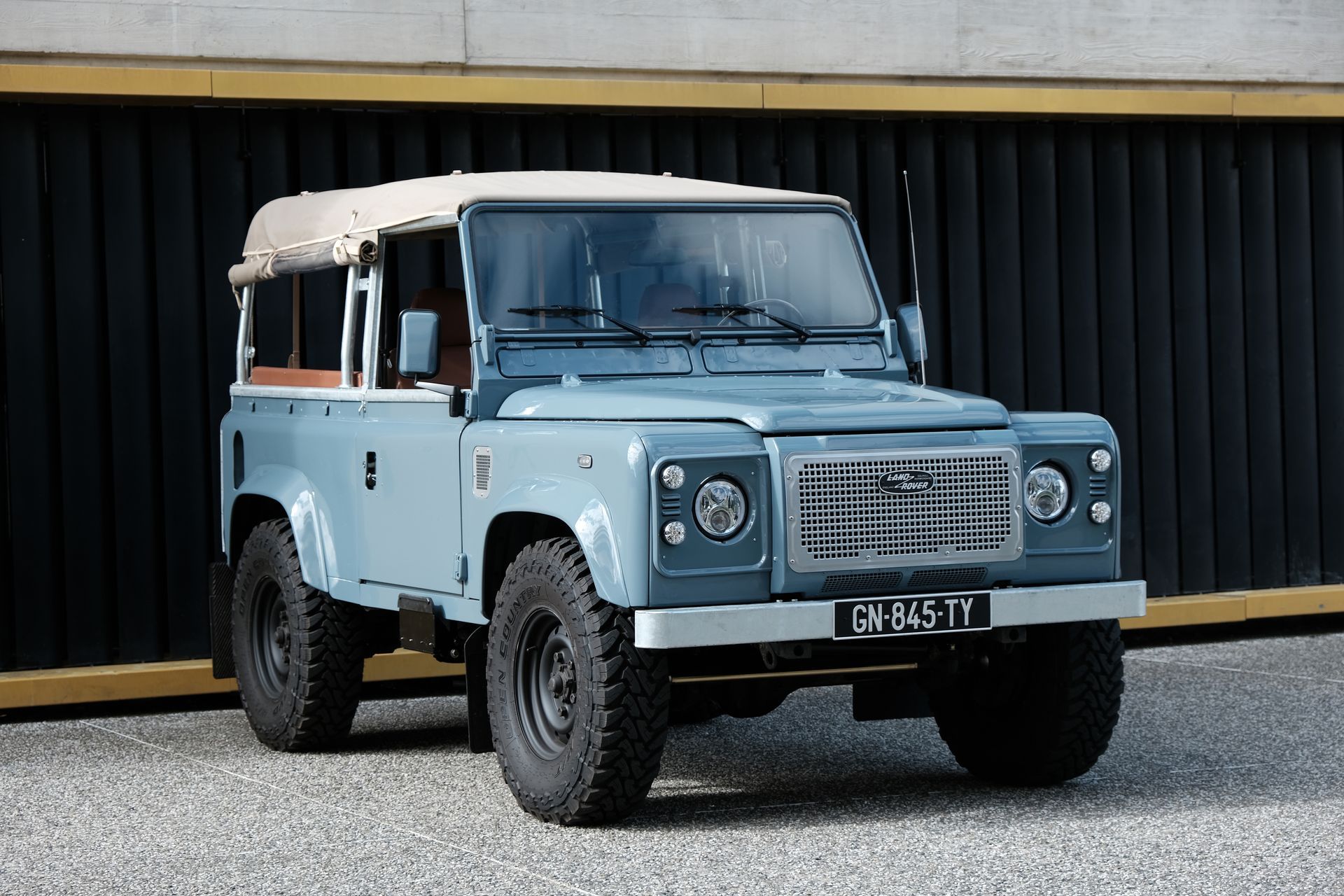 Un 4x4 Land Rover Defender bleu avec un toit couleur crème, garé sur du gravier devant un bâtiment sombre.