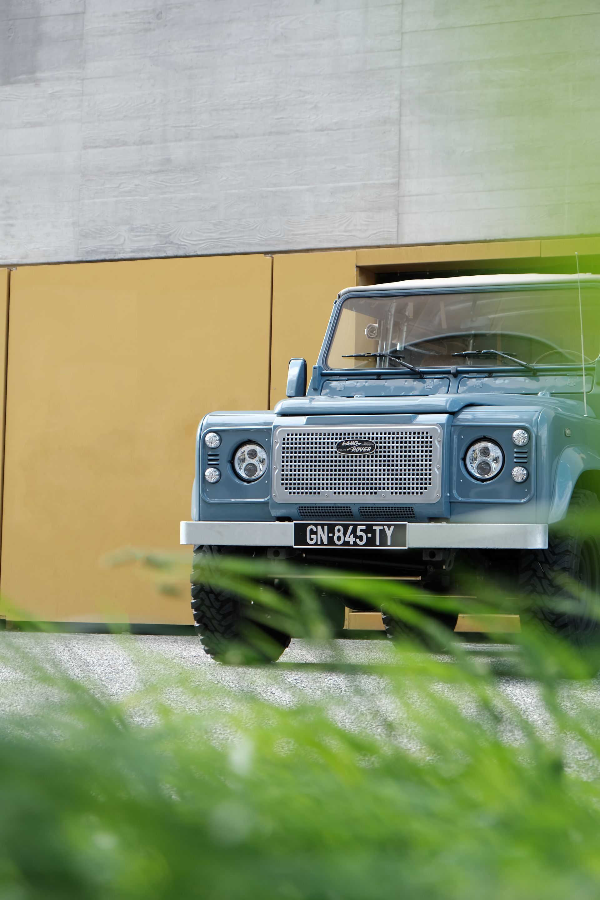 Un Land Rover Defender bleu est garé contre un mur jaune et gris, avec de hautes herbes au premier plan.