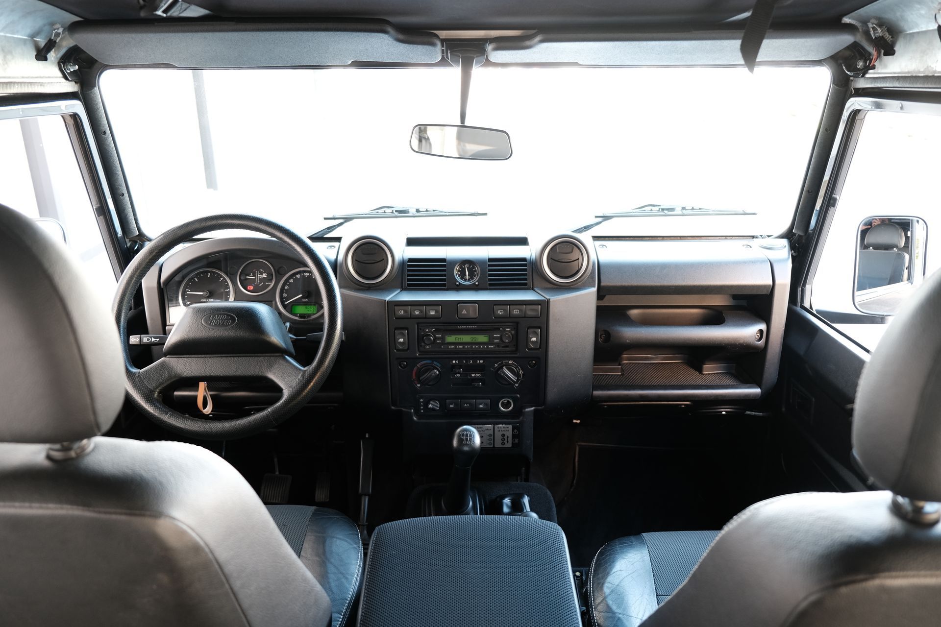 Vue intérieure d'un Land Rover Defender : tableau de bord noir, volant, sièges et radio avec console centrale.