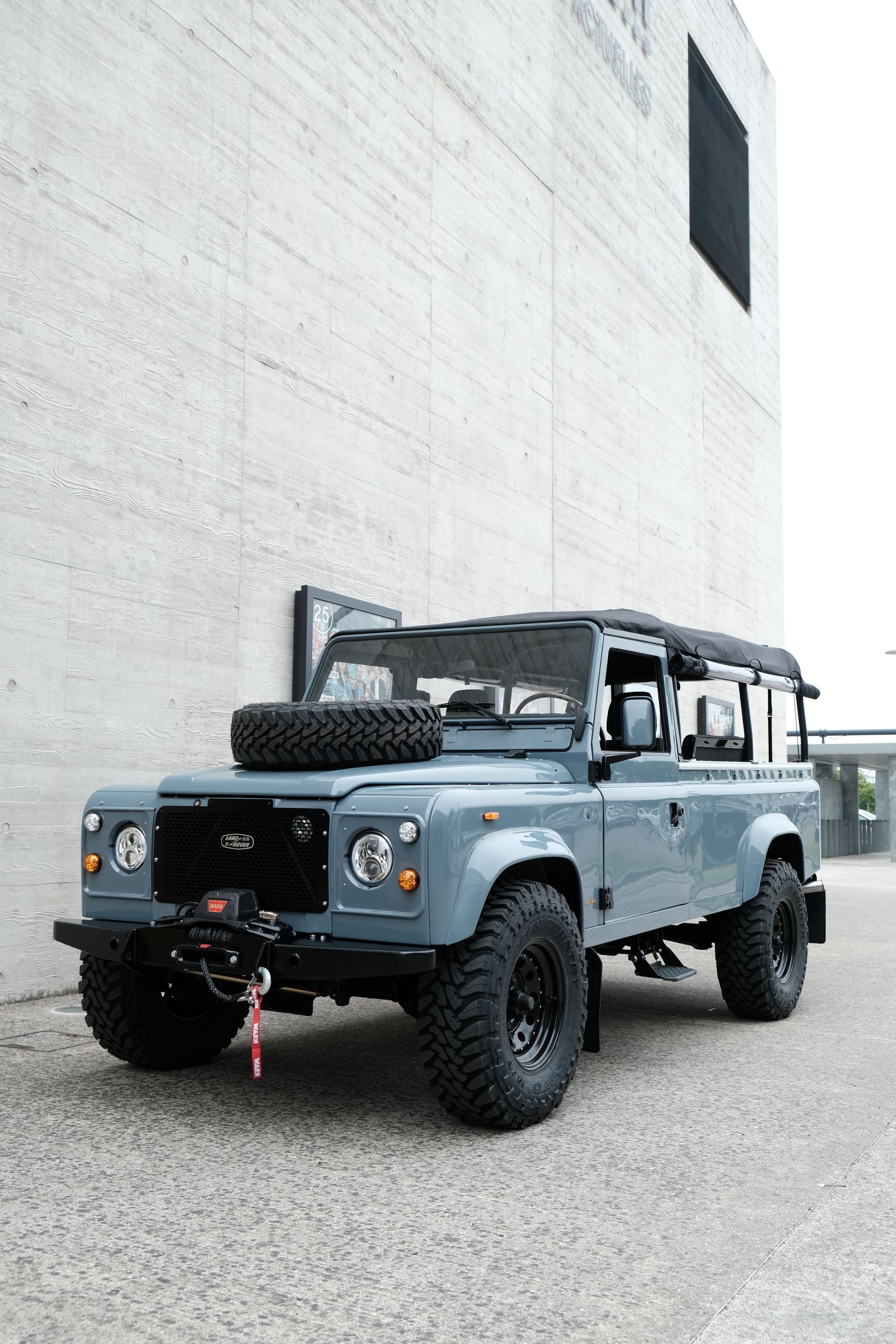 Land Rover Defender bleu garé devant un bâtiment en béton. Jantes noires, roue de secours sur le capot.