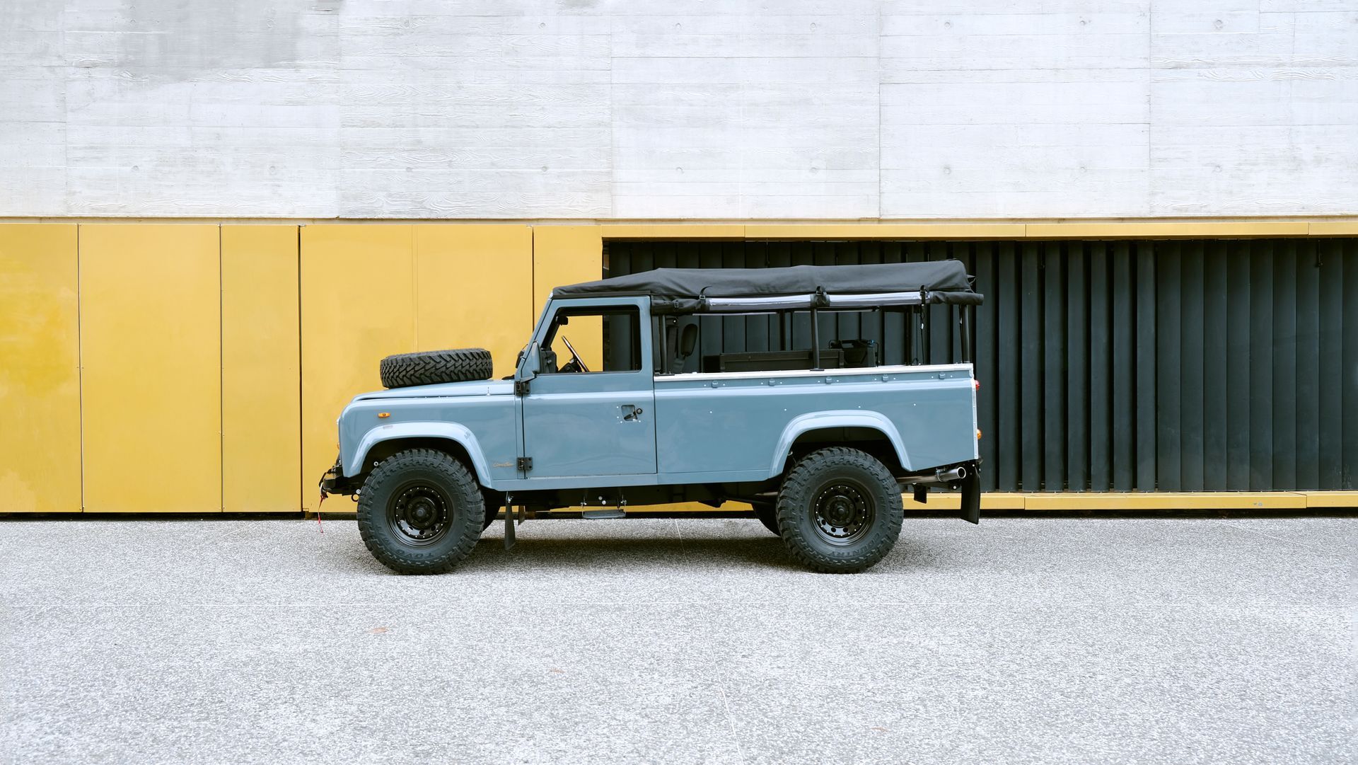 Un Land Rover Defender bleu est garé devant un bâtiment en béton aux panneaux jaunes.