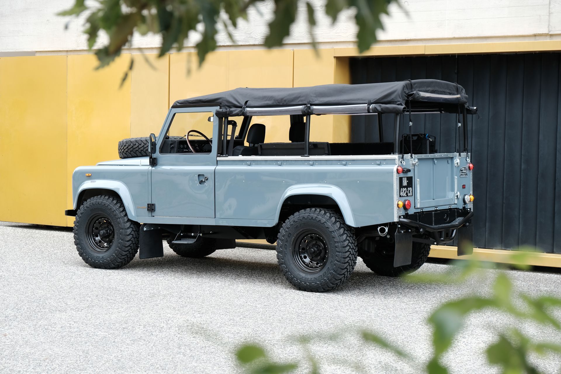 Un Land Rover Defender gris est garé à l'extérieur, avec une capote souple noire et des pneus tout-terrain.