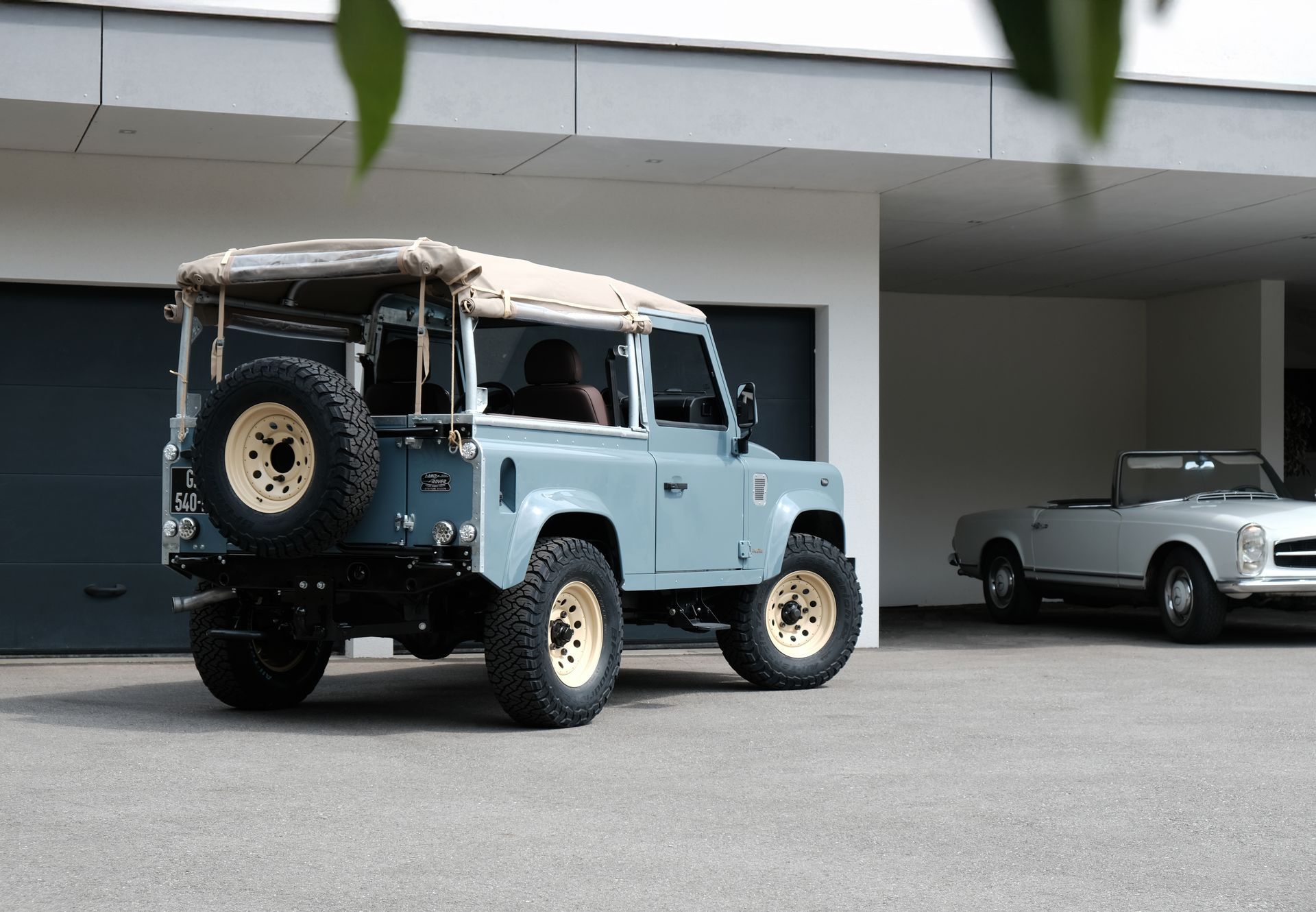 Un Land Rover Defender bleu clair avec un toit et des jantes beiges est garé dans une allée à côté d'un cabriolet argenté.