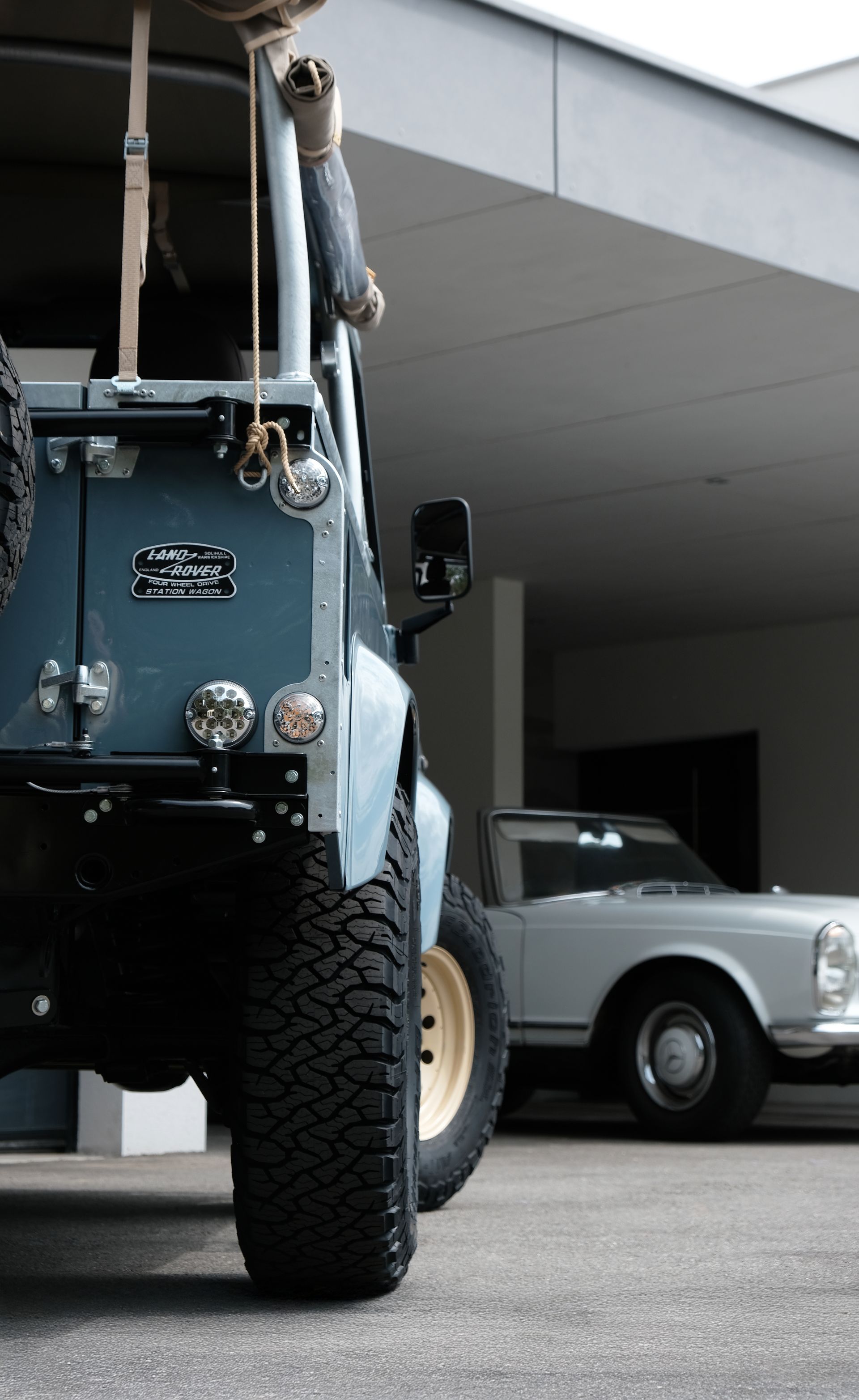 Un véhicule tout-terrain bleu clair à gros pneus est garé à côté d'une voiture ancienne blanche sous un auvent.