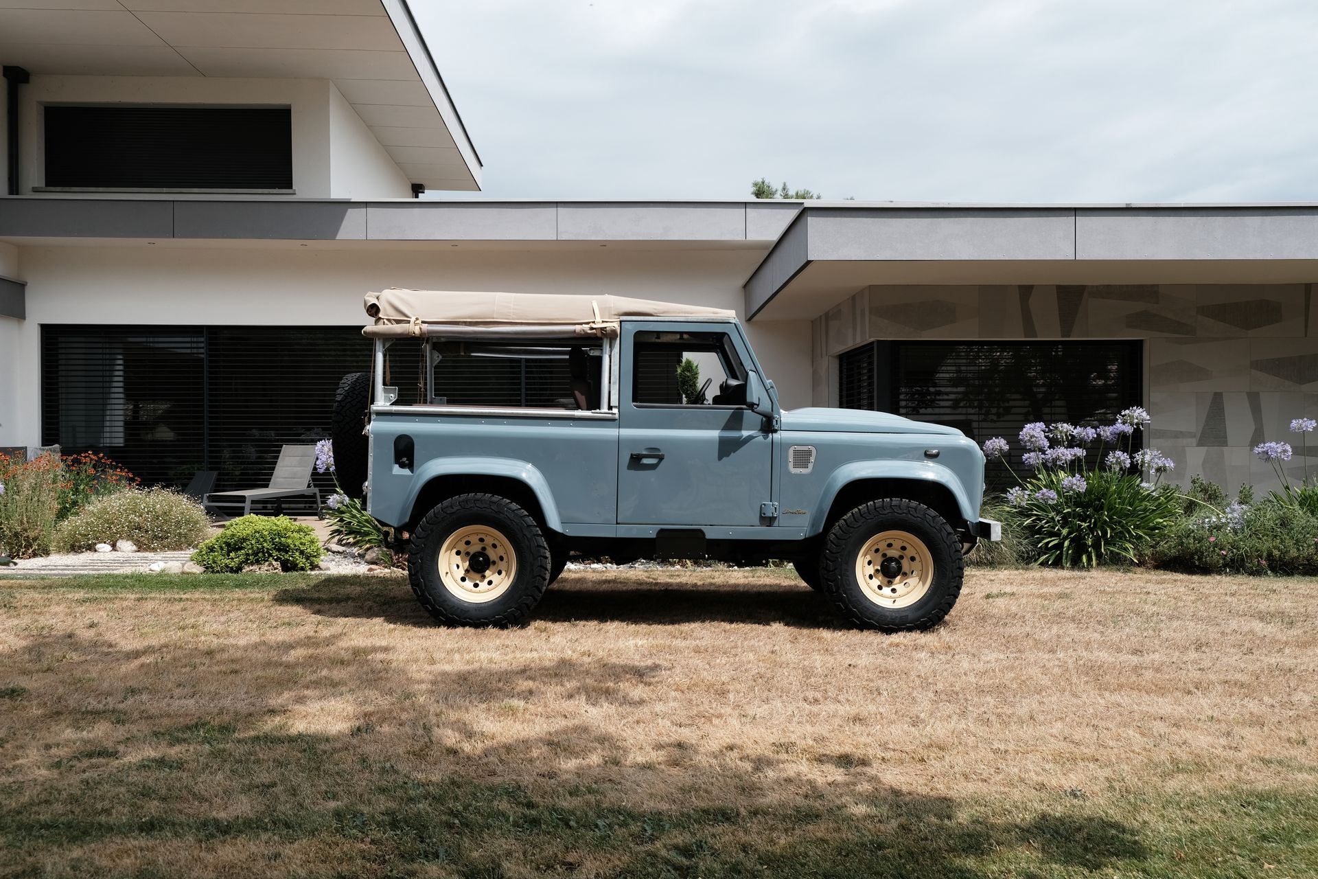 Un Land Rover Defender bleu clair, avec toit beige et jantes dorées, est garé devant une maison moderne.
