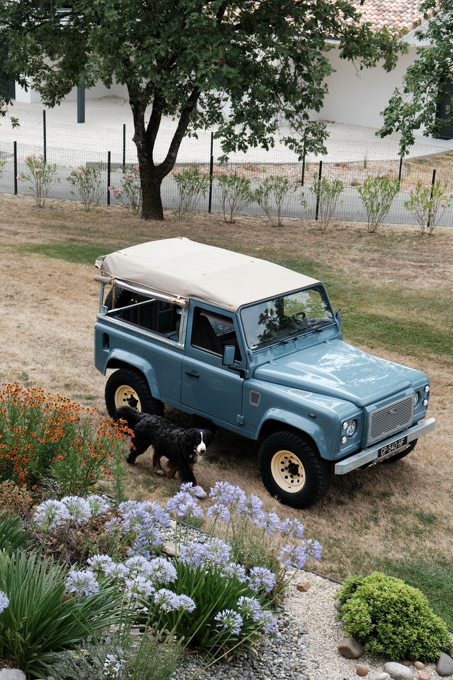 Un Land Rover Defender bleu clair avec des jantes dorées est garé devant une maison blanche moderne par une journée ensoleillée.