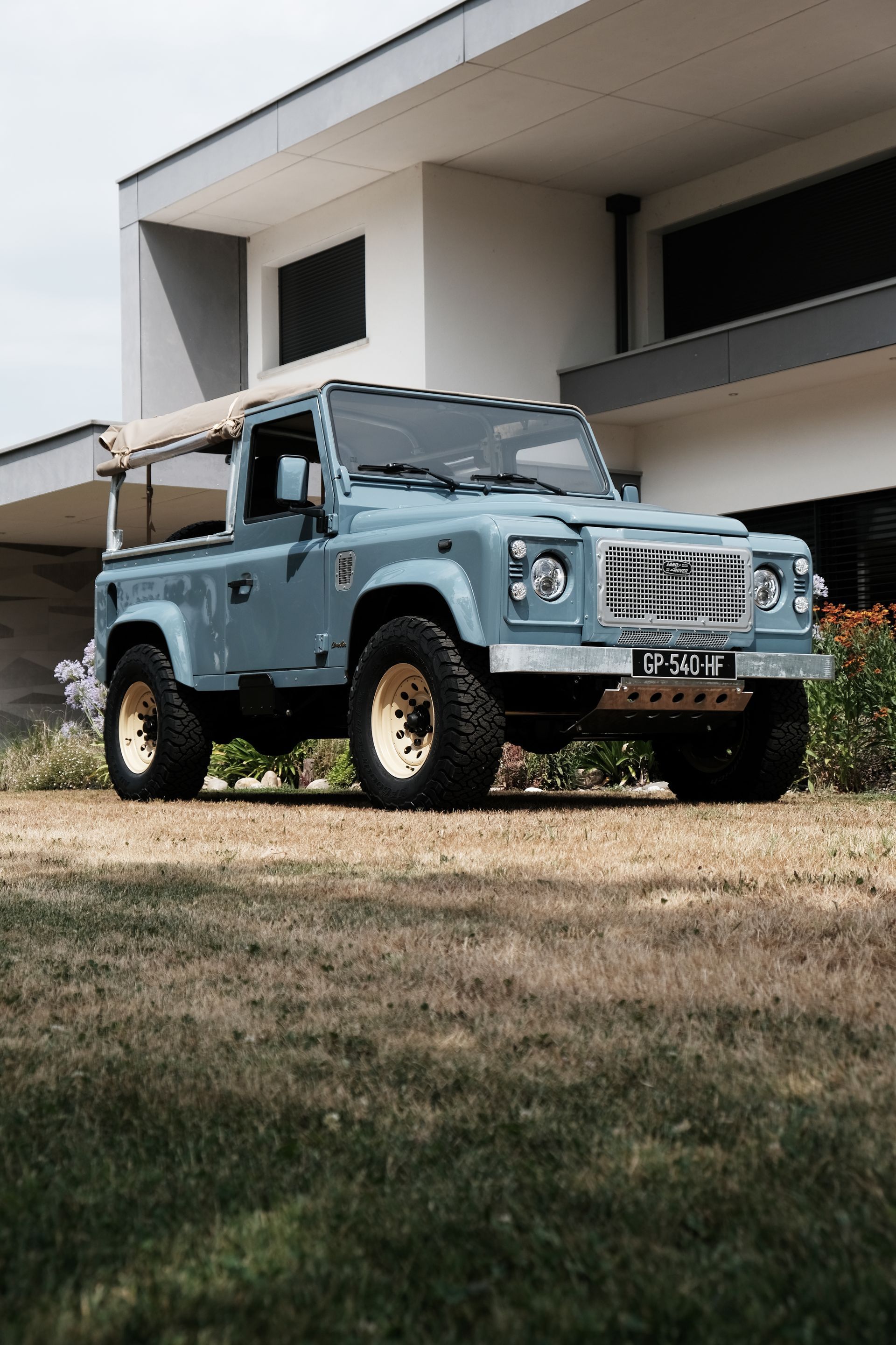 Un Land Rover Defender bleu clair avec des jantes dorées est garé devant un bâtiment blanc moderne.