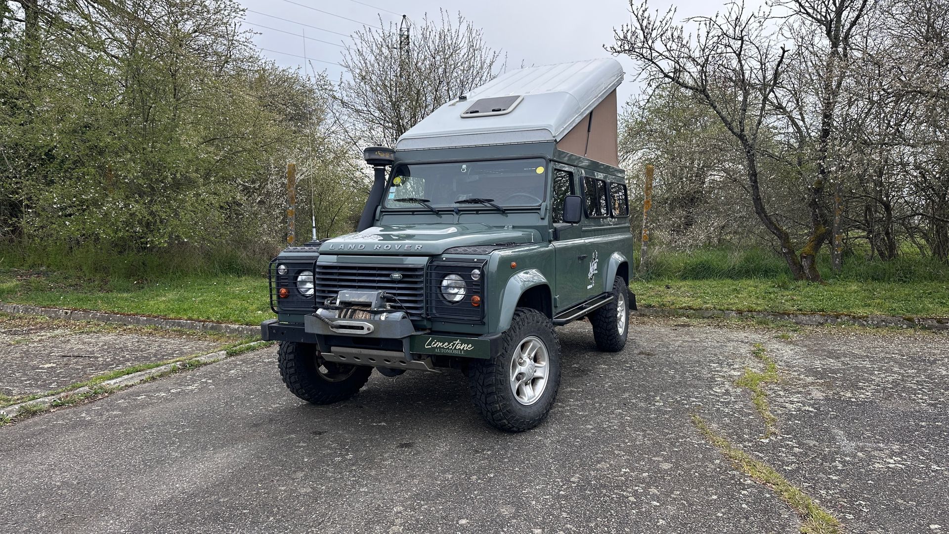 Land Rover Defender verte avec une tente de toit sur une surface pavée près des arbres.