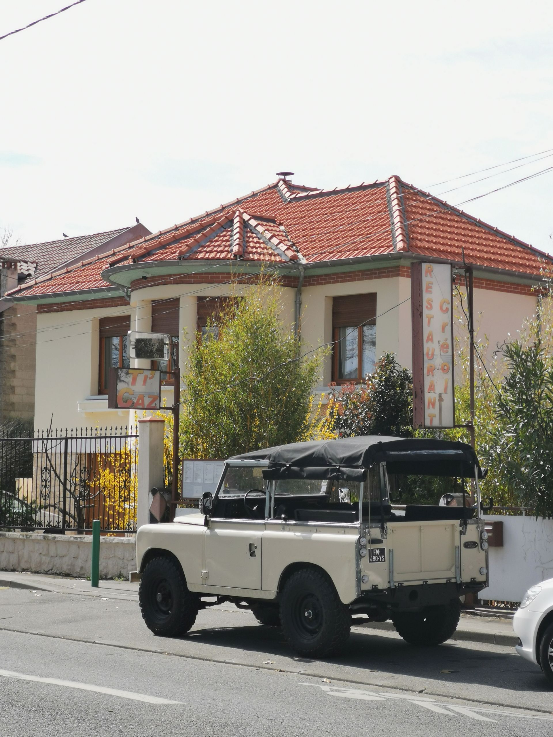 Un Land Rover couleur crème est garé devant un bâtiment jaune clair au toit de tuiles rouges.